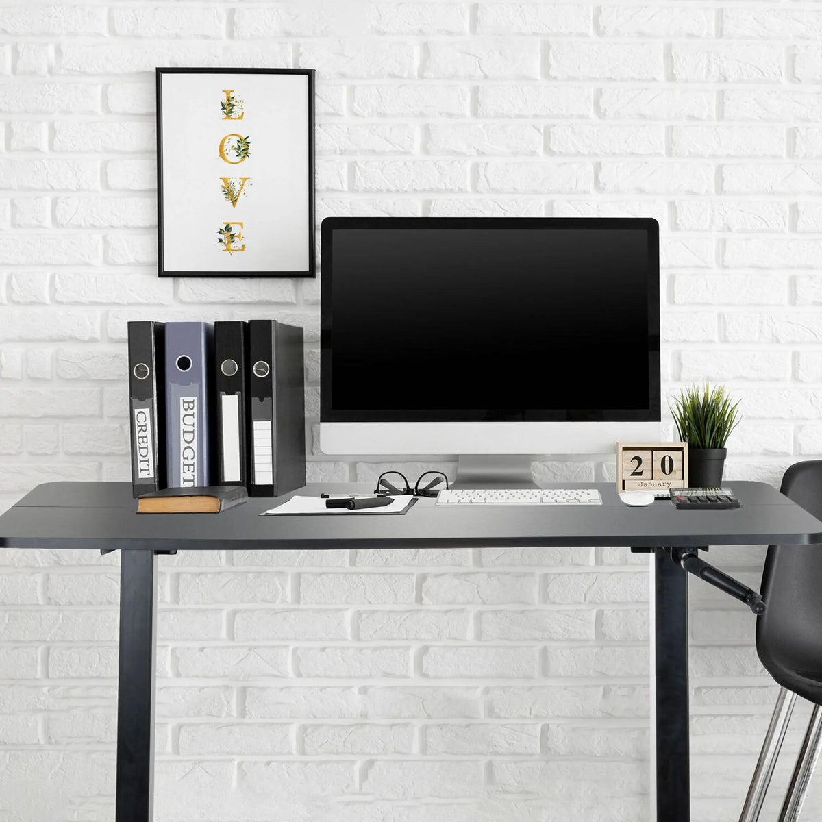 Musta seisomatyöpöytä STAND CR näytöllä, näppäimistöllä ja kansioilla valkoista tiiliseinää vasten, valmistaja hjh OFFICE